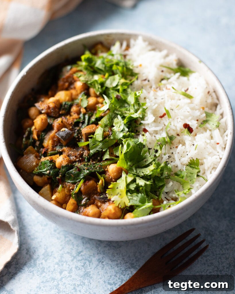 26 Cozy Vegan Dinners to Warm Your Winter Nights 5 Light blue ceramic bowl filled with chickpea, potato, eggplant and spinach curry with gravy and rice to the side with green cilantro garnish in the center. The bowl is on a gray tabletop with a light yellow and white striped cloth napkin to the left and a dark brown wood serving in the front to the right