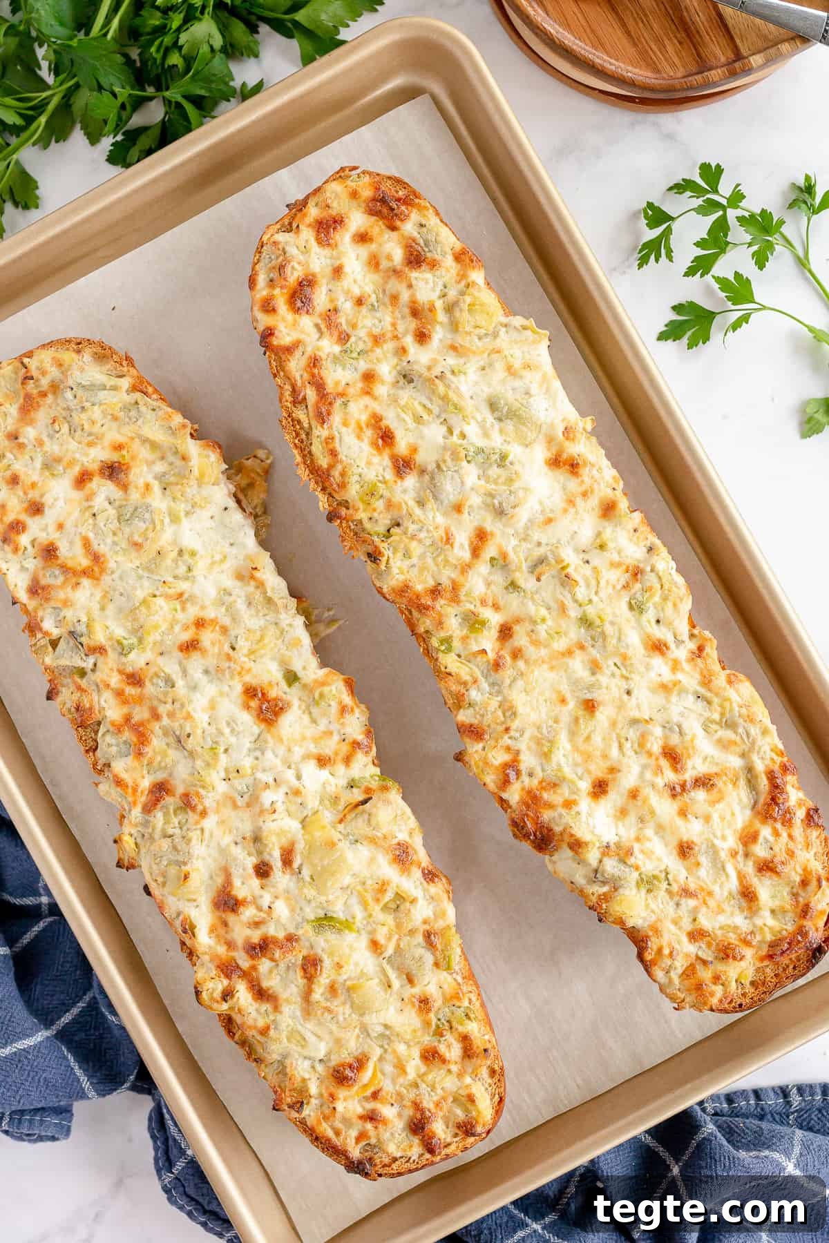 Cheesy Artichoke Pull-Apart Bread for Parties and Snacks 3 Two halves of a loaf of French bread topped with a browned cheese artichoke mixture on a baking sheet.