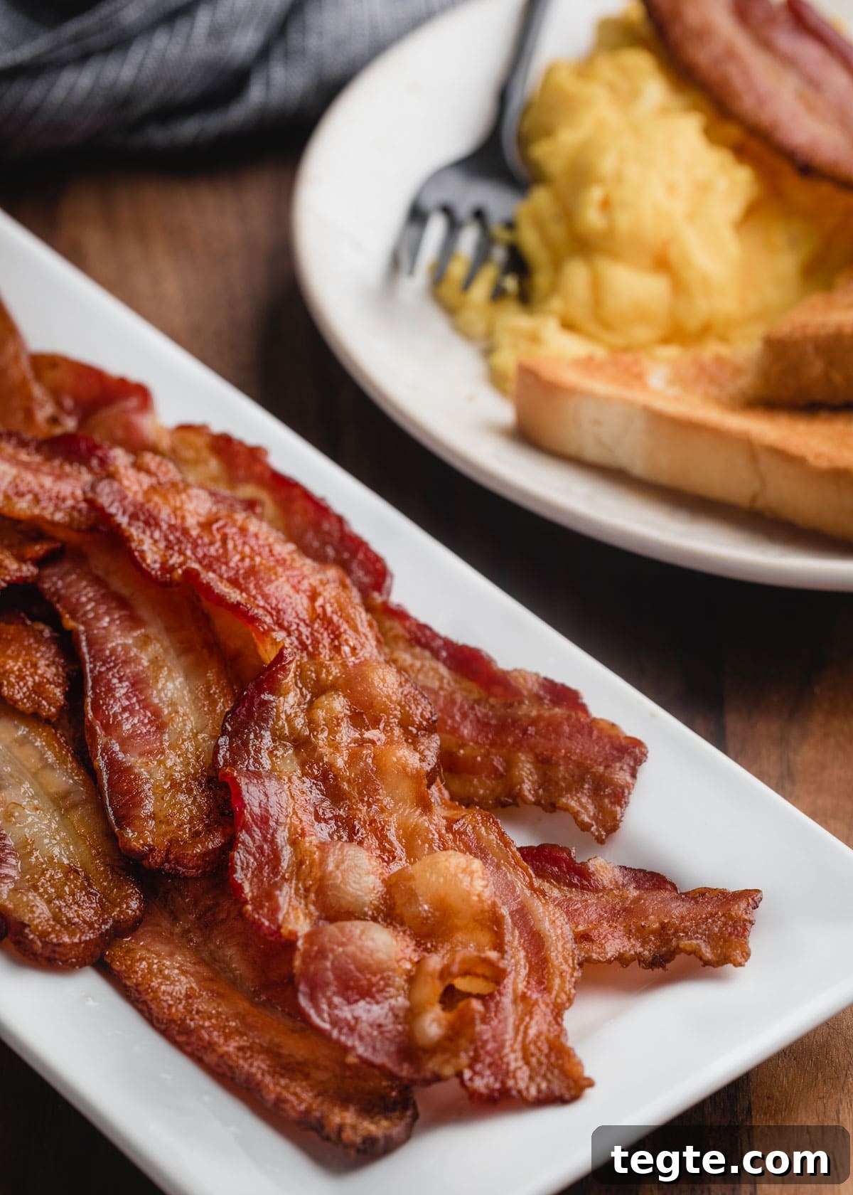 baked bacon on a white platter in front of a breakfast plate of scrambled eggs and toast