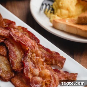 baked bacon on a white platter in front of a breakfast plate of scrambled eggs and toast