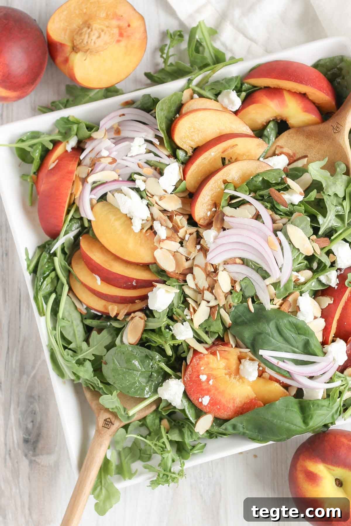 Wooden salad tongs mixing together a peach and goat cheese salad on a square plate with peaches on the sides.