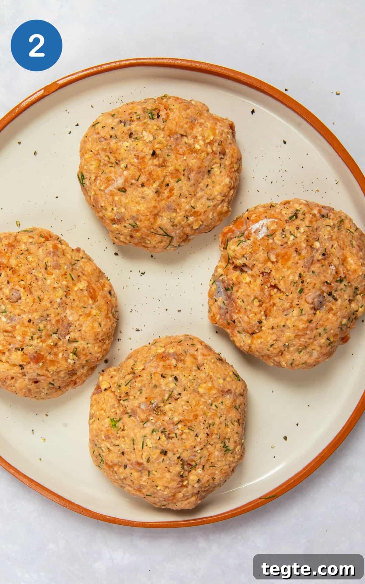 Crispy Salmon Burgers with Lemon-Dill Sauce 5 uncooked salmon burgers in patty shapes on white plate with orange rim.