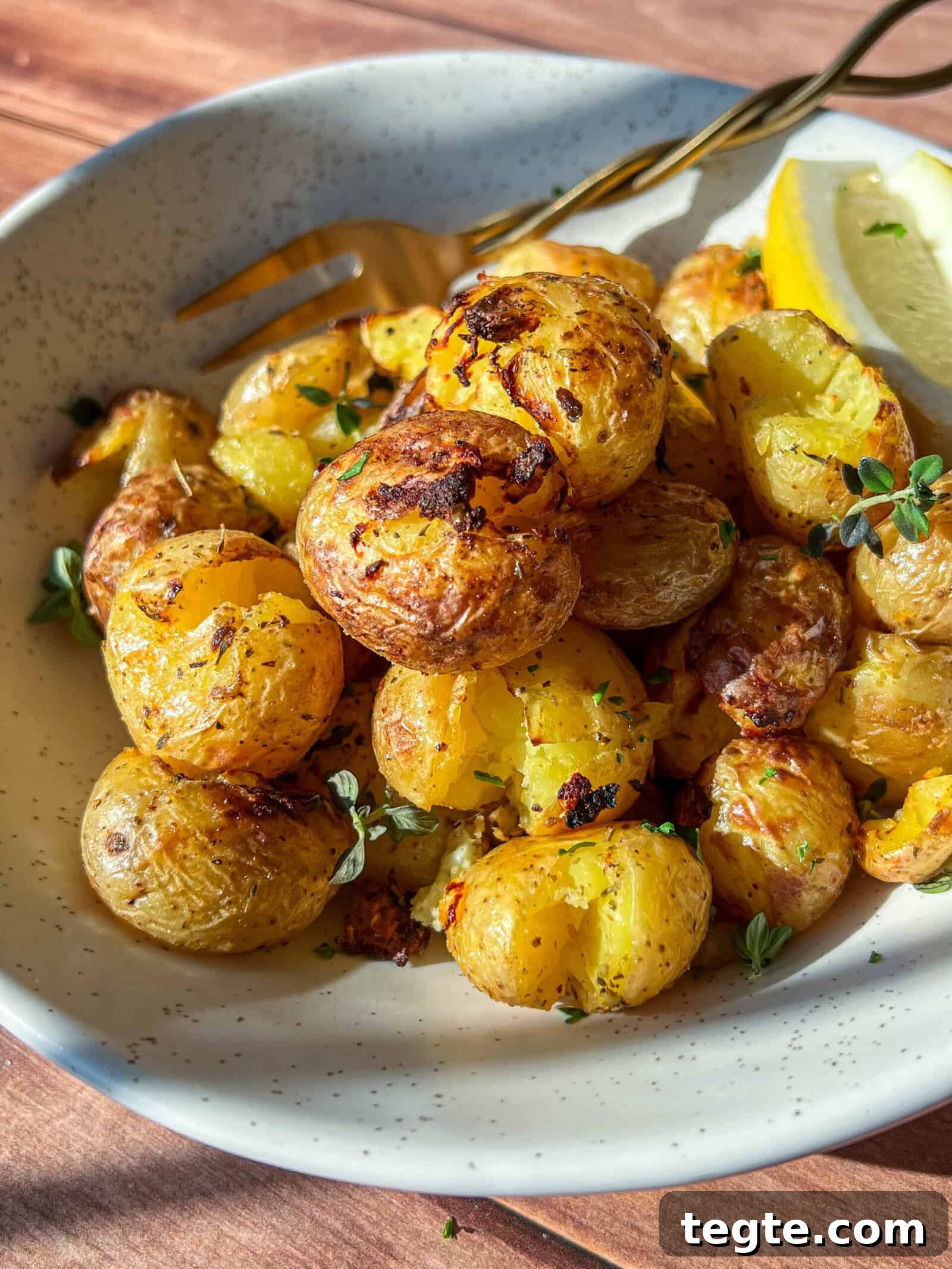 Crispy Greek Lemon Potatoes in the Air Fryer 2 A bowl of crispy roasted baby potatoes garnished with fresh herbs, served with a lemon wedge and a fork on the side, all on a speckled ceramic plate.