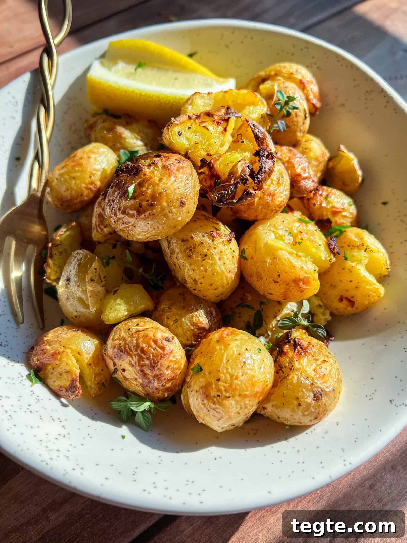 Crispy Greek Lemon Potatoes in the Air Fryer 3 A white speckled bowl filled with roasted baby potatoes garnished with herbs and two lemon wedges, with a gold fork resting on the edge, on a wooden surface.
