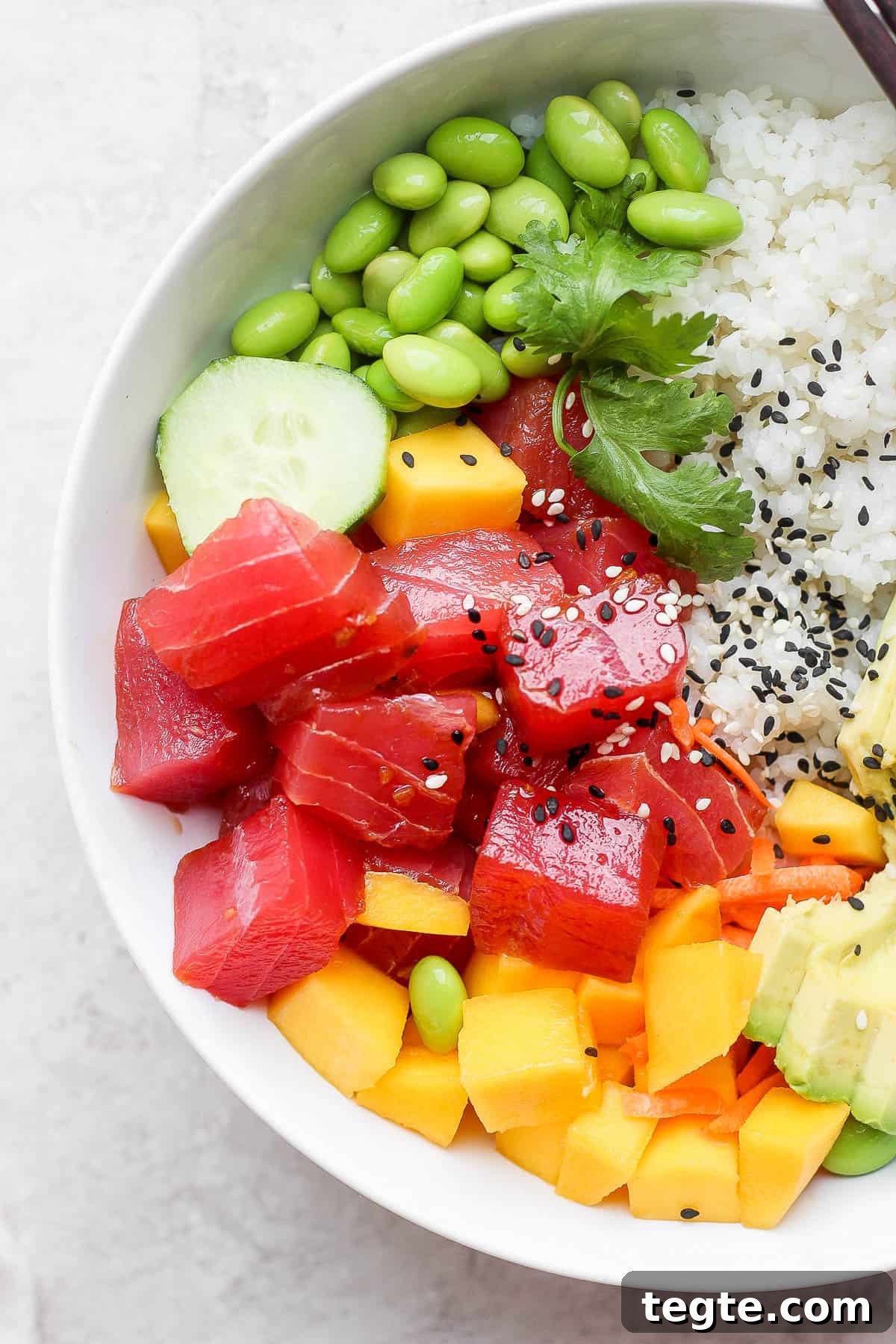 Fresh Tuna Poke Bowl Recipe: Quick Hawaiian-Style Lunch 7 Tuna poke bowl with sushi rice, edamame, cucumber, mango, avocado, cilantro, and garnished with sesame seeds.
