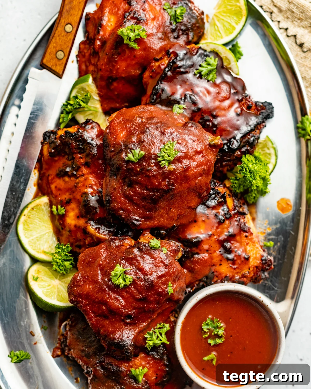 Oven-Baked BBQ Chicken Thighs with Crispy Skin and Sticky Glaze 8 bbq chicken thighs on a plate with a knife and a bowl of bbq sauce.
