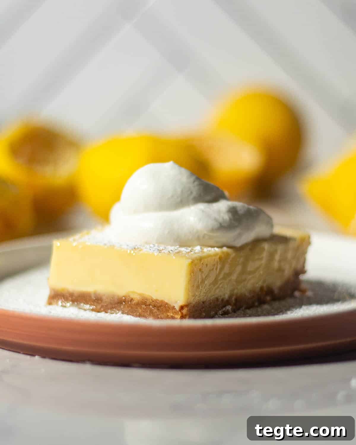 A single lemon bar with a graham cracker crust on a white plate, topped with whipped cream and lightly dusted with powdered sugar, surrounded by halved lemons.