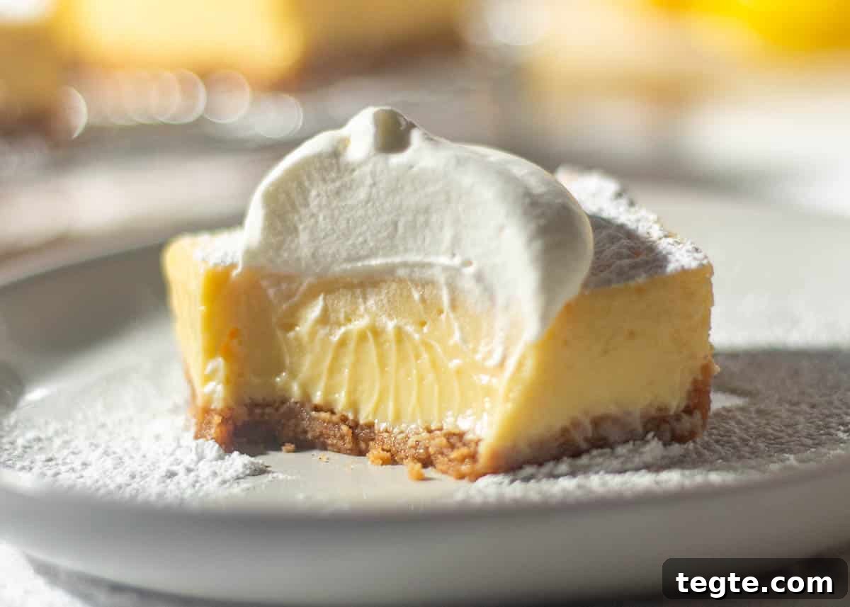 A close-up of a half-eaten lemon bar with a graham cracker crust, revealing the creamy lemon center and topped with whipped cream, with a blurred background of more lemon bars.