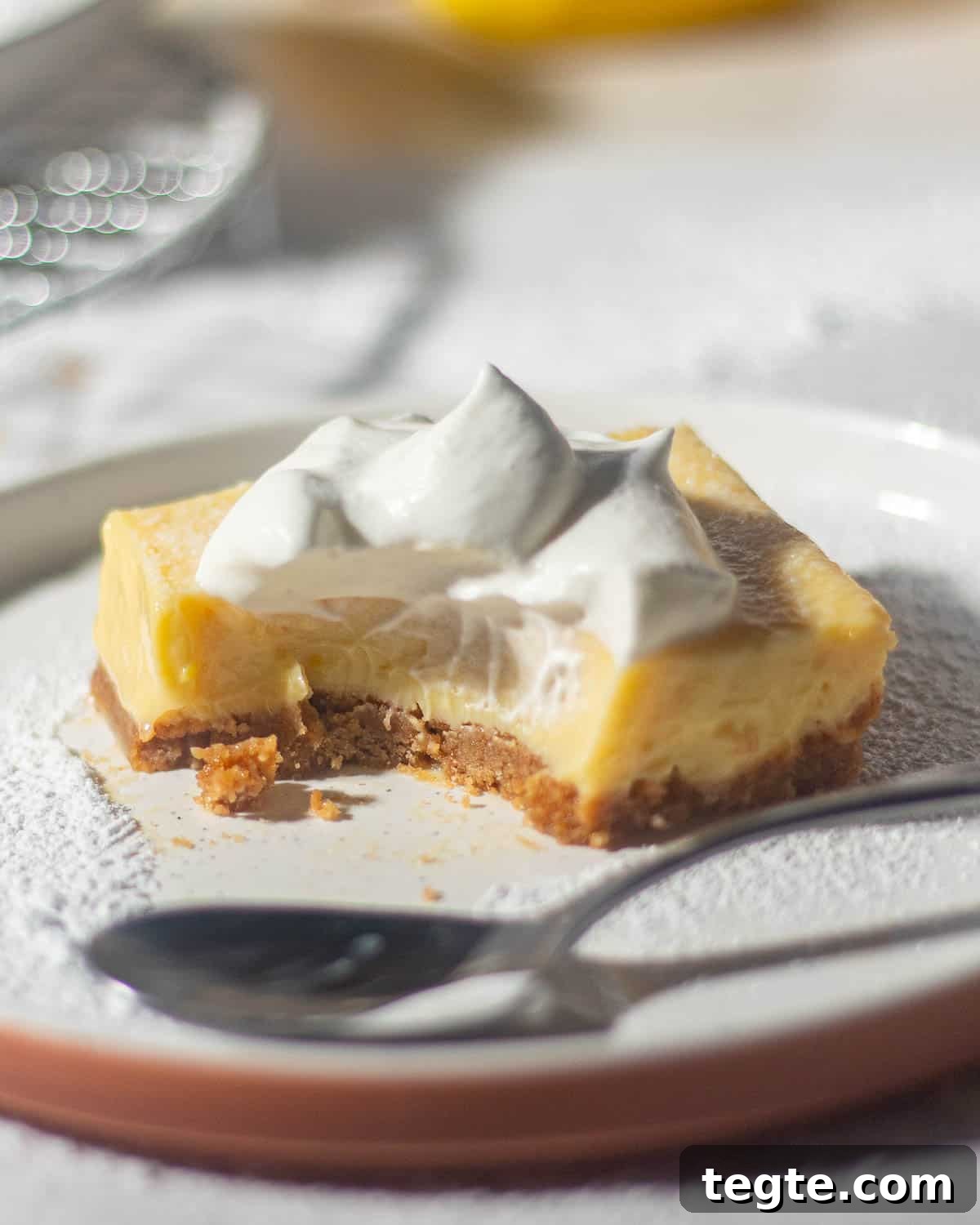 A partially eaten lemon bar with a graham cracker crust and whipped cream on top, served on a white plate with a spoon next to it, showing the creamy lemon filling.