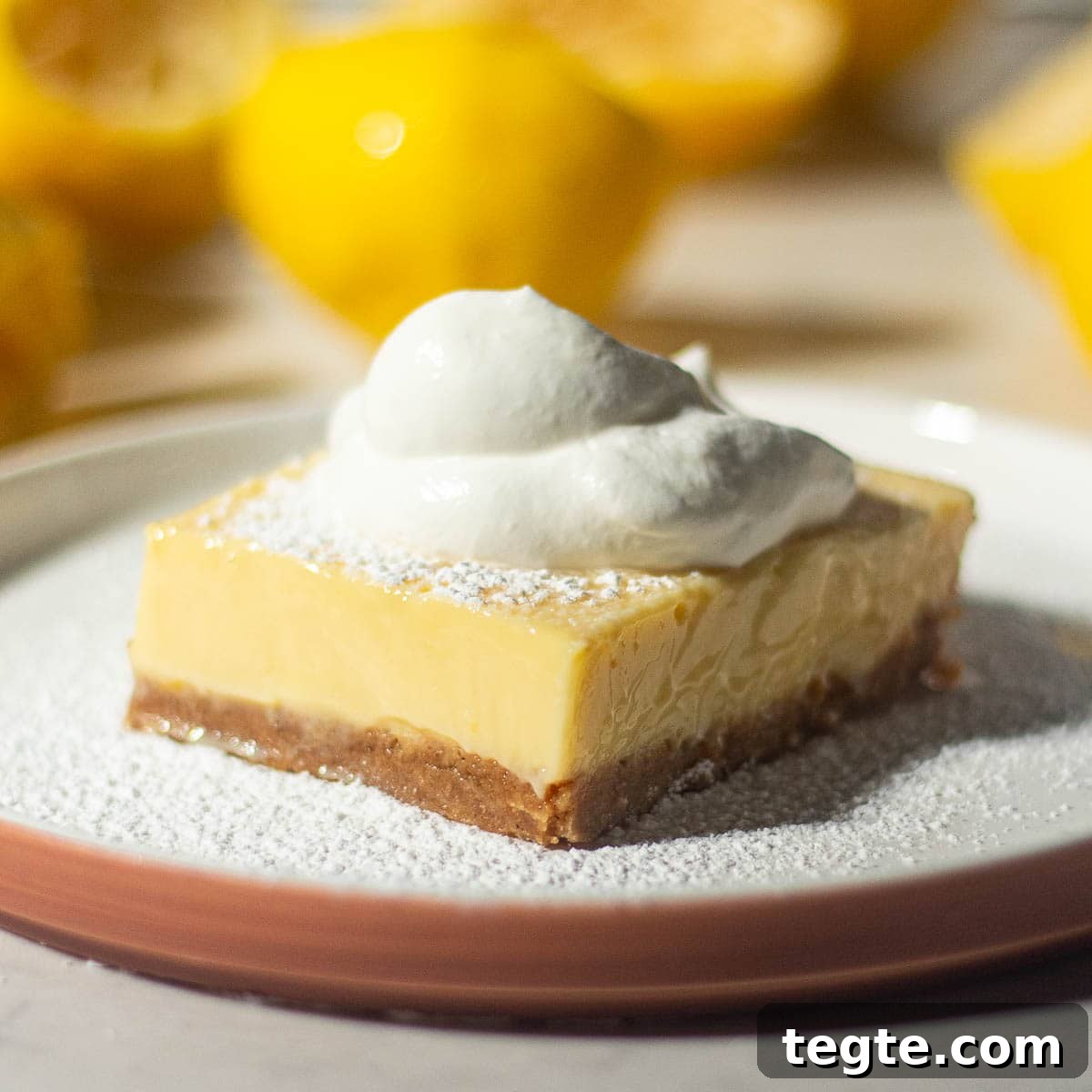 A single lemon bar with a graham cracker crust on a white plate, topped with whipped cream and lightly dusted with powdered sugar, surrounded by halved lemons.