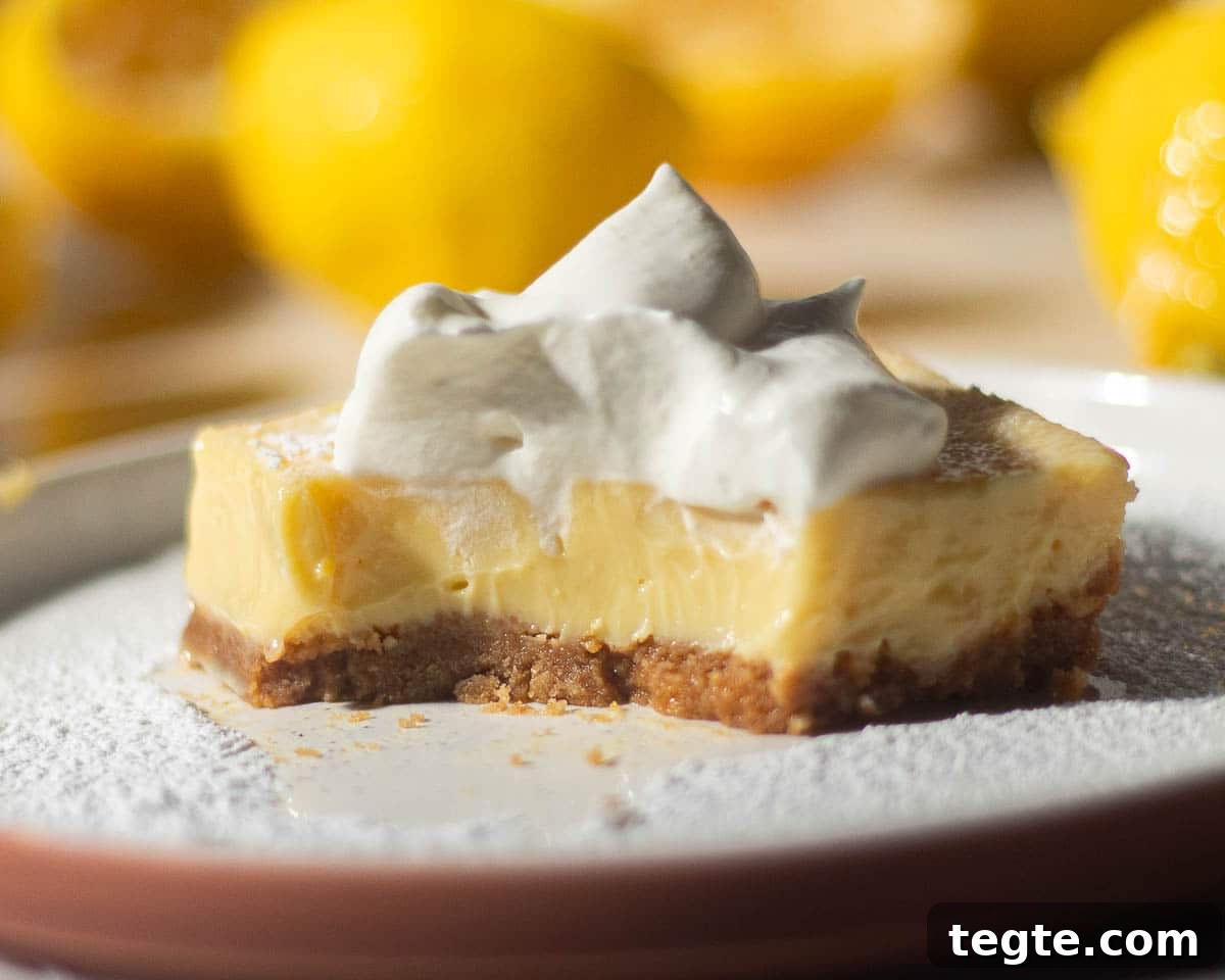 A partially eaten lemon bar with a graham cracker crust and whipped cream on top, revealing the creamy lemon filling, with lemons blurred in the background.