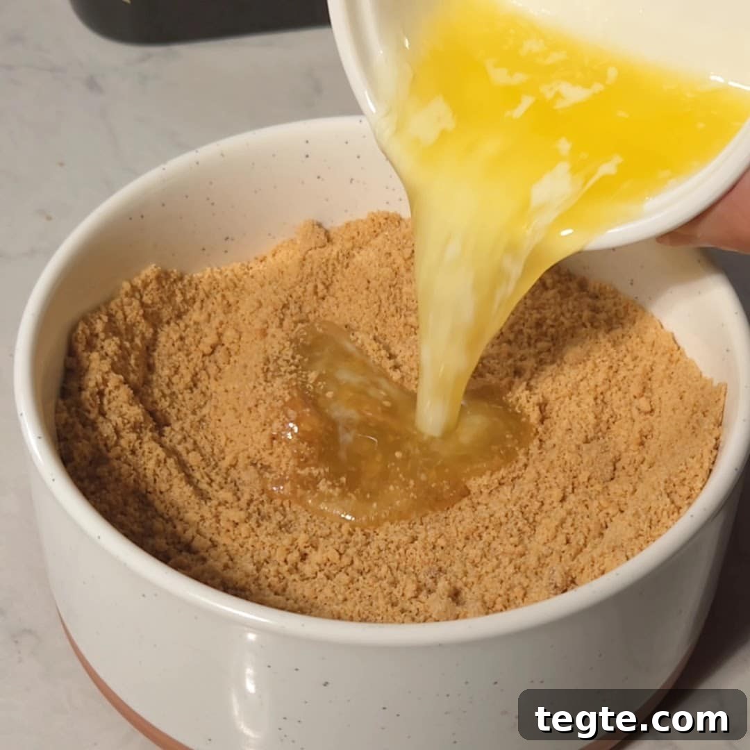 Melted butter being poured into a bowl of graham cracker crumbs to create the crust for lemon bars, with a smooth and consistent mixture being formed.
