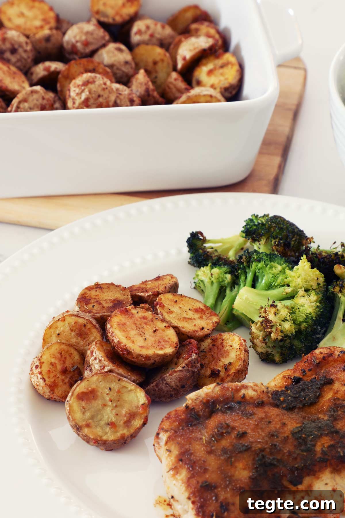 Crispy Air-Fryer Baby Potatoes Recipe 2 A white plate with air fryer baby potatoes, broccoli and pork chops with a baking dish full of potatoes on a kitchen counter.