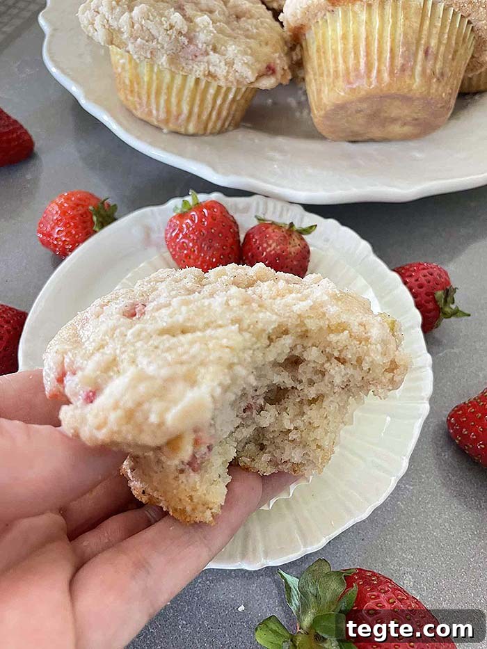 A strawberry rhubarb muffin with a bite out of it held above a small plate with two whole strawberries. Whole strawberries are scattered around the small plate and a partial picture of a plate bowl with strawberry rhubarb muffins is at the top.