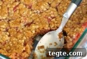 A glass 8x8 dish of rhubarb crisp with a serving taken out of a corner with a serving spoon in its place. The text Rhubarb Crisp is overlaid near the top of the photo. The text reallifedinner.com is overlaid at the bottom.