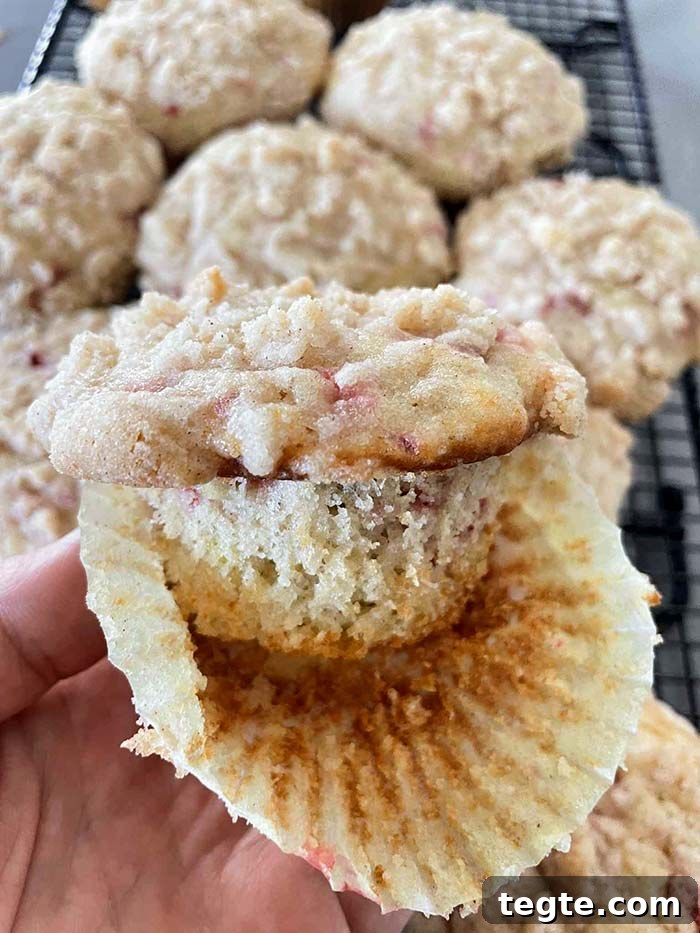 A strawberry rhubarb muffin with the paper liner peeled halfway off held above a cooling rack with strawberry rhubarb muffins.