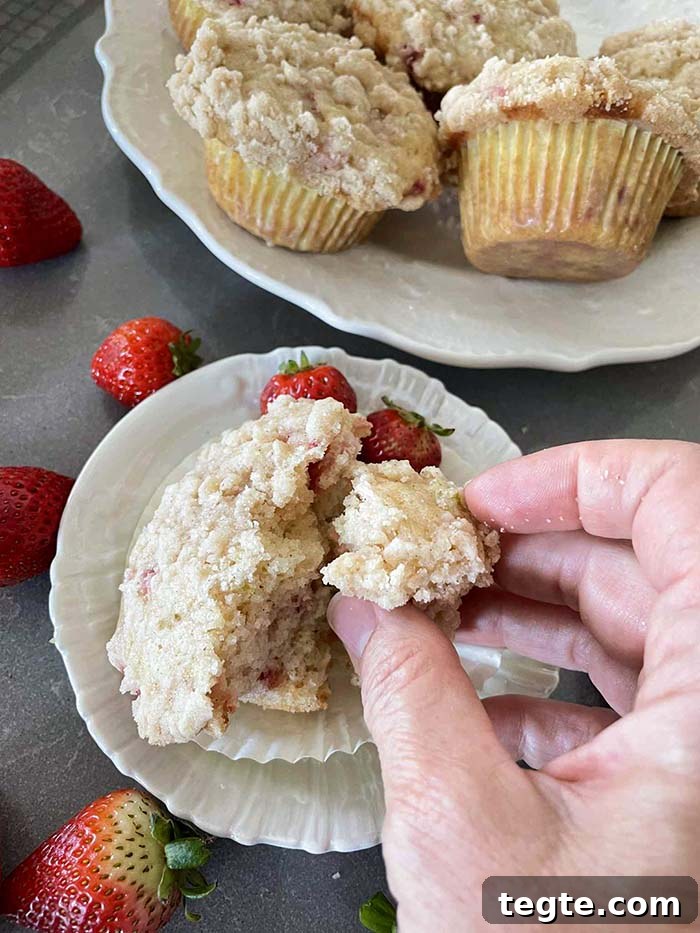 A half of strawberry rhubarb muffin in a peeled paper liner on a small plate with a hand breaking off a piece of muffin. Whole strawberries are scattered around the small plate. In the upper right corner is a plate bowl of strawberry rhubarb muffins.
