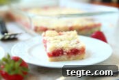 Faded in the background is a serving spatula, a whole strawberry and a glass 9x13 dish of strawberry rhubarb crumb bars with a piece cut out of the corner. In front of the baking dish is a square piece of strawberry rhubarb bar on a dessert plate with a fork handle in front and a whole strawberry in the left corner.