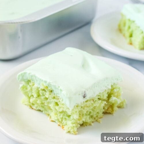 slice of pistachio pineapple cake topped with whipped cream frosting, served on a white plate. The pan of cake and another slice are in the background.