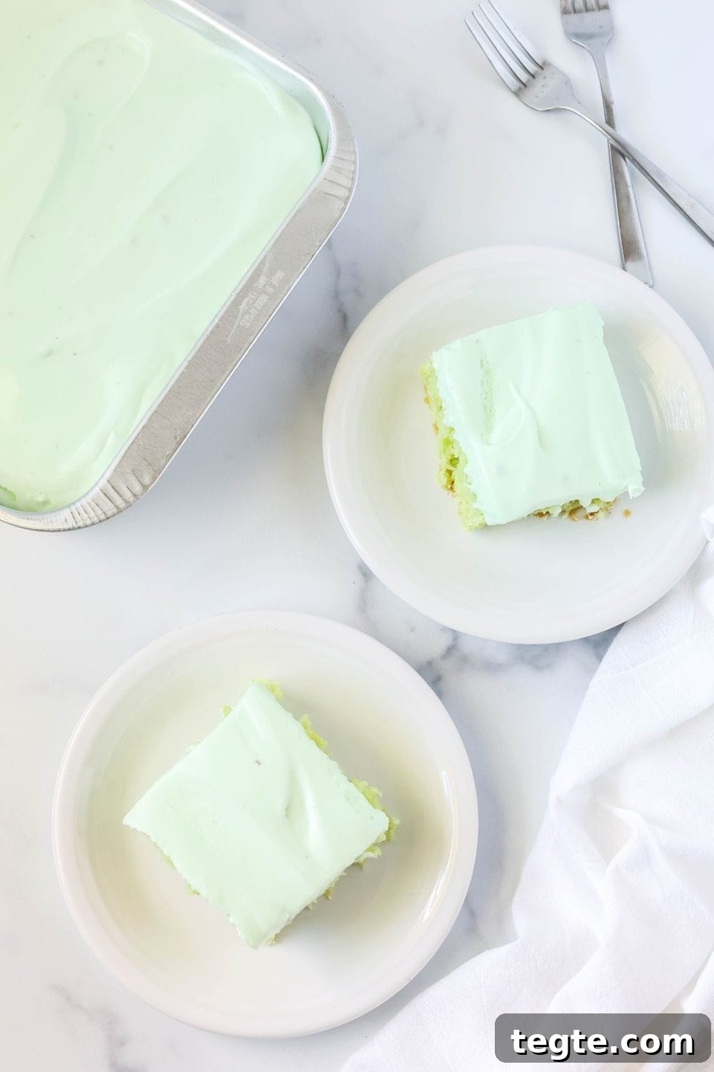 overhead view of two white plates serving pieces of pineapple pistachio cake. The pan of remaining cake is next to the plates, along with two forks.