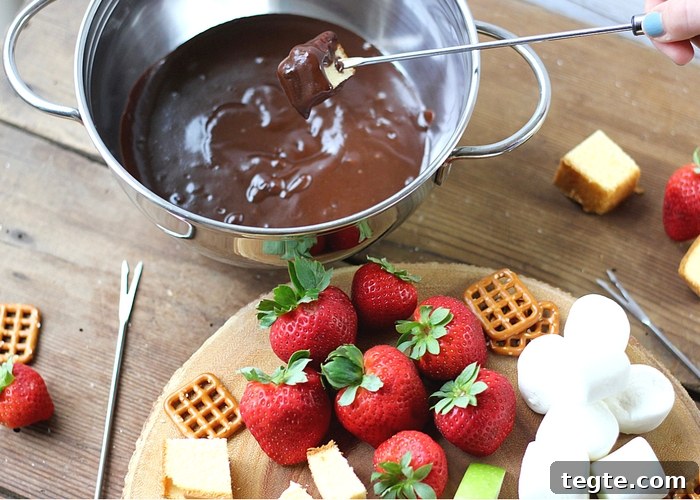 Chocolate Caramel Fondue Recipe for Dipping Fruits and Treats 3 A pot full of chocolate caramel fondue with a piece of cake dipping into it. Fresh fruit, marshmallows and pretzels are on a platter next to it.