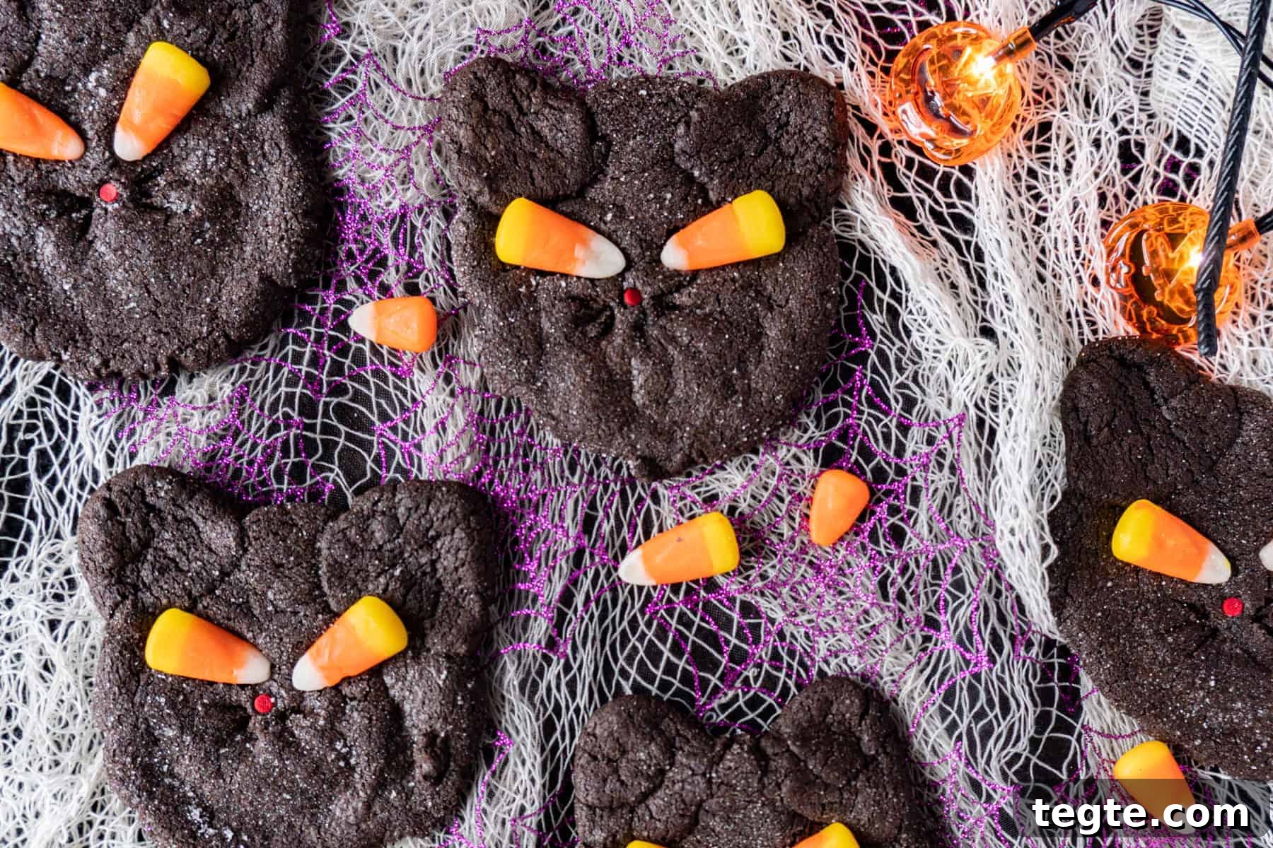 Black Cat Cookie Recipe: Soft Chocolate Drops for Halloween 4 Black Cat Cookies on a purple and white cheesecloth background surrounded by candy corn pieces and pumpkin lights.
