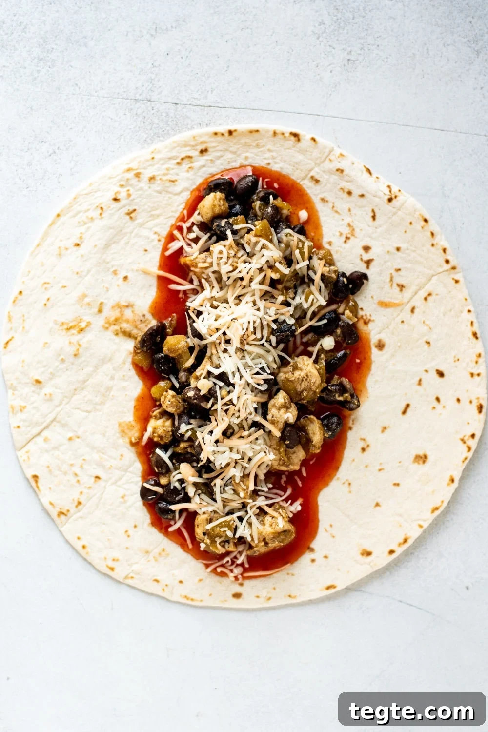 An uncooked burrito tortilla lying flat on a surface topped with seasoned chicken, black beans, red enchilada sauce, and Mexican blend cheese.