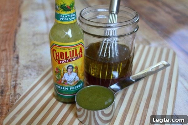 Making dressing for salad with a bottle of cholulua green hot sauce, a measuring cup and a jar with a whisk. set on a cutting board.