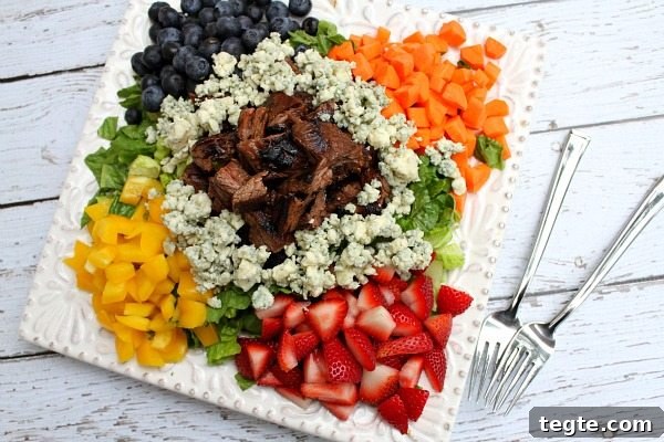Grilled Steak Chopped Summer Salad on a square white plate with steak piled in the middle and veggies, lettuce and fruit and blue cheese crumbles piled around the steak. two forks on the side. on a white plank table