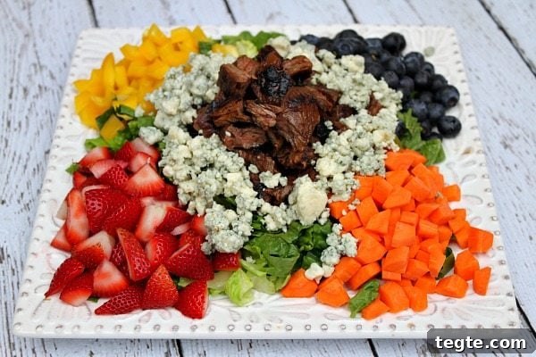 Grilled Steak Chopped Summer Salad on a square white plate with steak piled in the middle and veggies, lettuce and fruit and blue cheese crumbles piled around the steak. on a white plank table