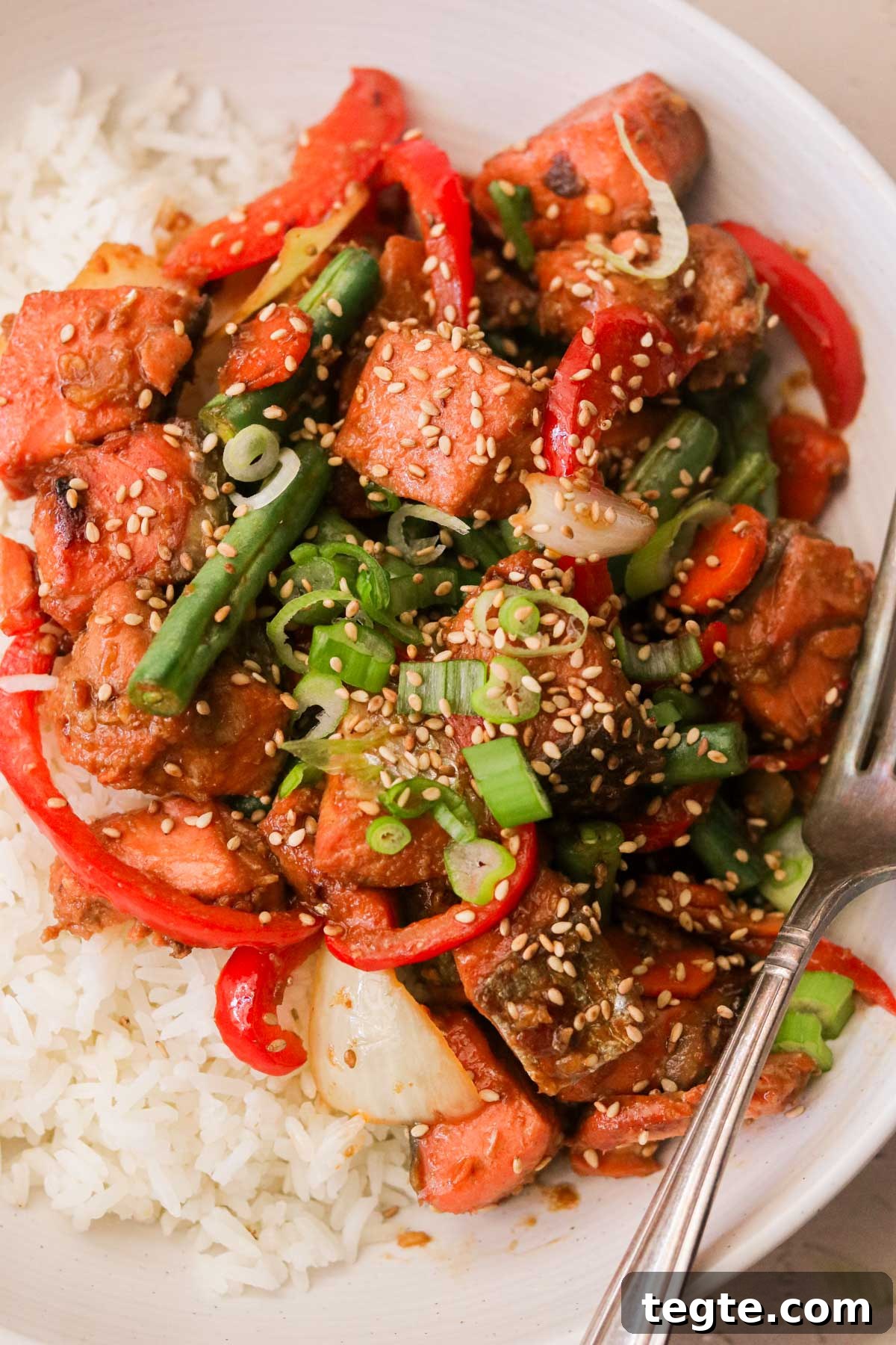 Quick Asian Salmon Stir-Fry with Garlic Soy Glaze 2 a close up image of salmon stir fry over white rice and garnished with green onions and sesame seeds with a silver fork resting next to it