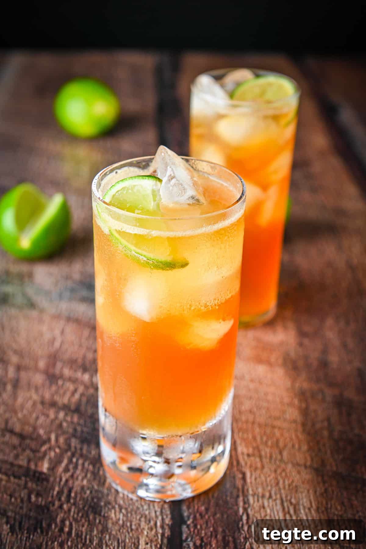 Two tall glasses with two dark and stormy cocktails in it along with limes on a table