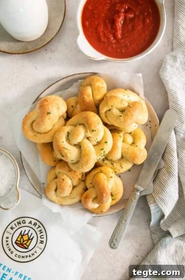 A plate of golden, twisted garlic knots topped with herbs sits on parchment paper next to a bowl of red marinara sauce, a butter knife, a striped napkin, and a King Arthur flour bag.