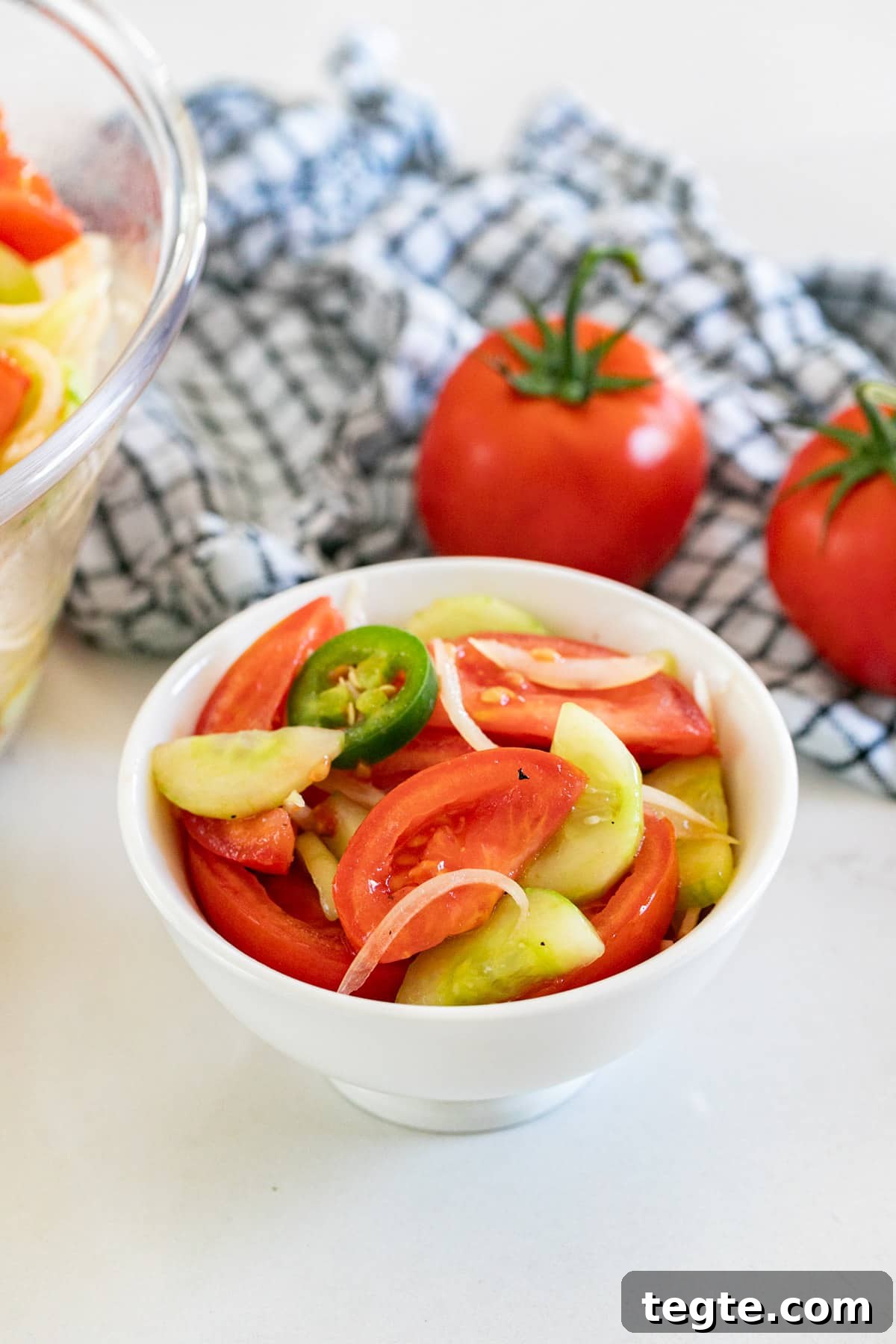A bowl of tomato salad with onions and cucumbers  with a checked towel and fresh tomatoes in the background.