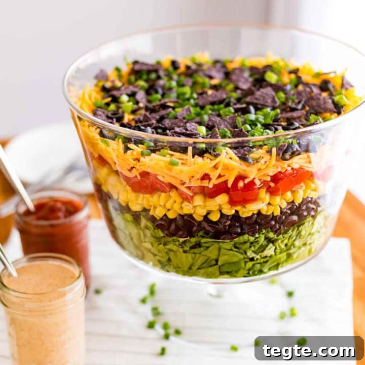 A side view of a clear glass trifle dish with a layered taco salad with the dressing and salsa in jars on the side.