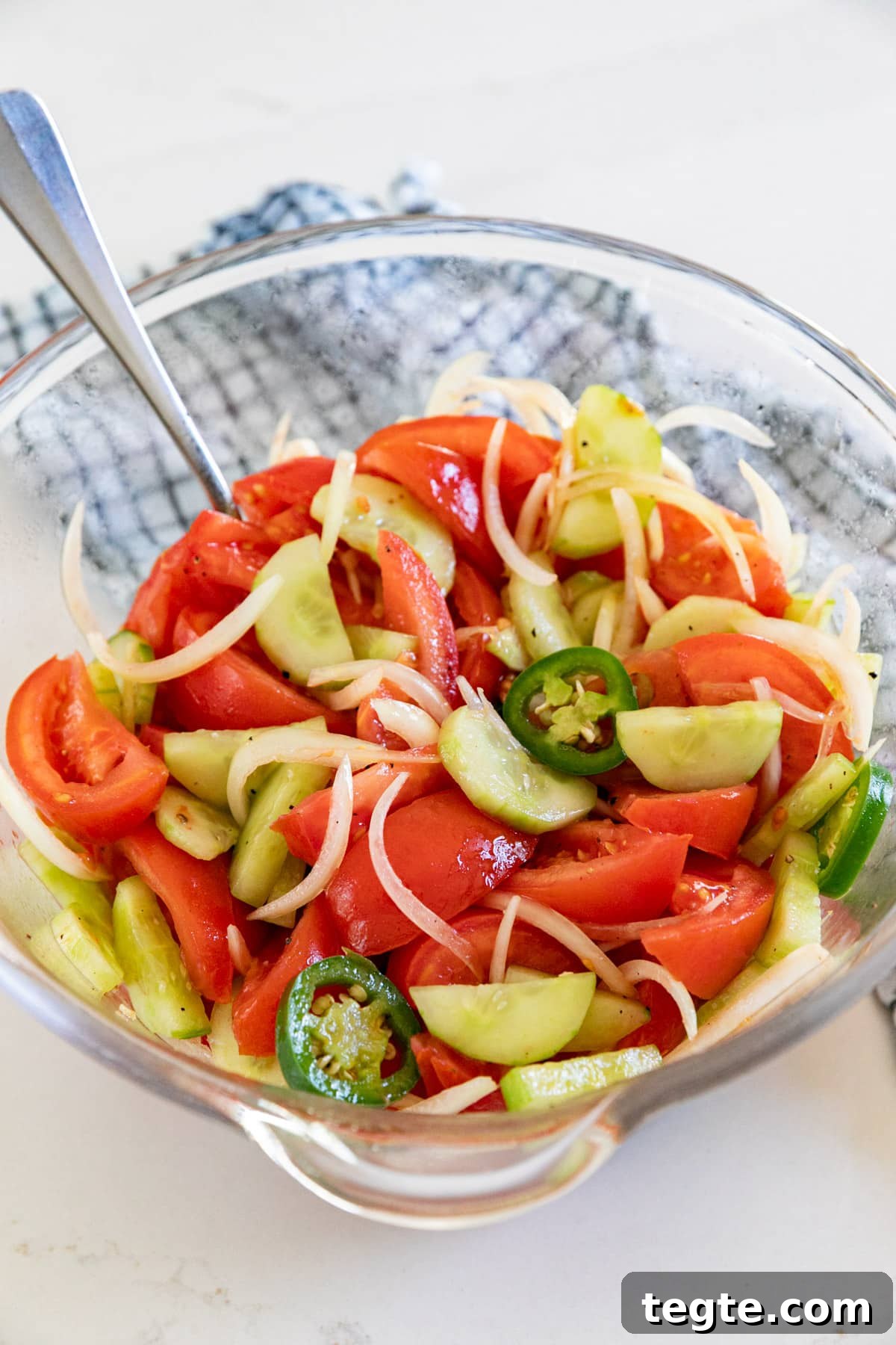 A large bowl of tomato salad with a spoon in it and a checked towel in the background.