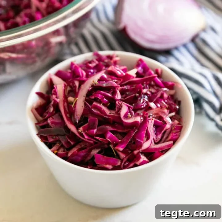 A white bowl of red cabbage salad with a large glass bowl of it and a chunk of red onion in the background.