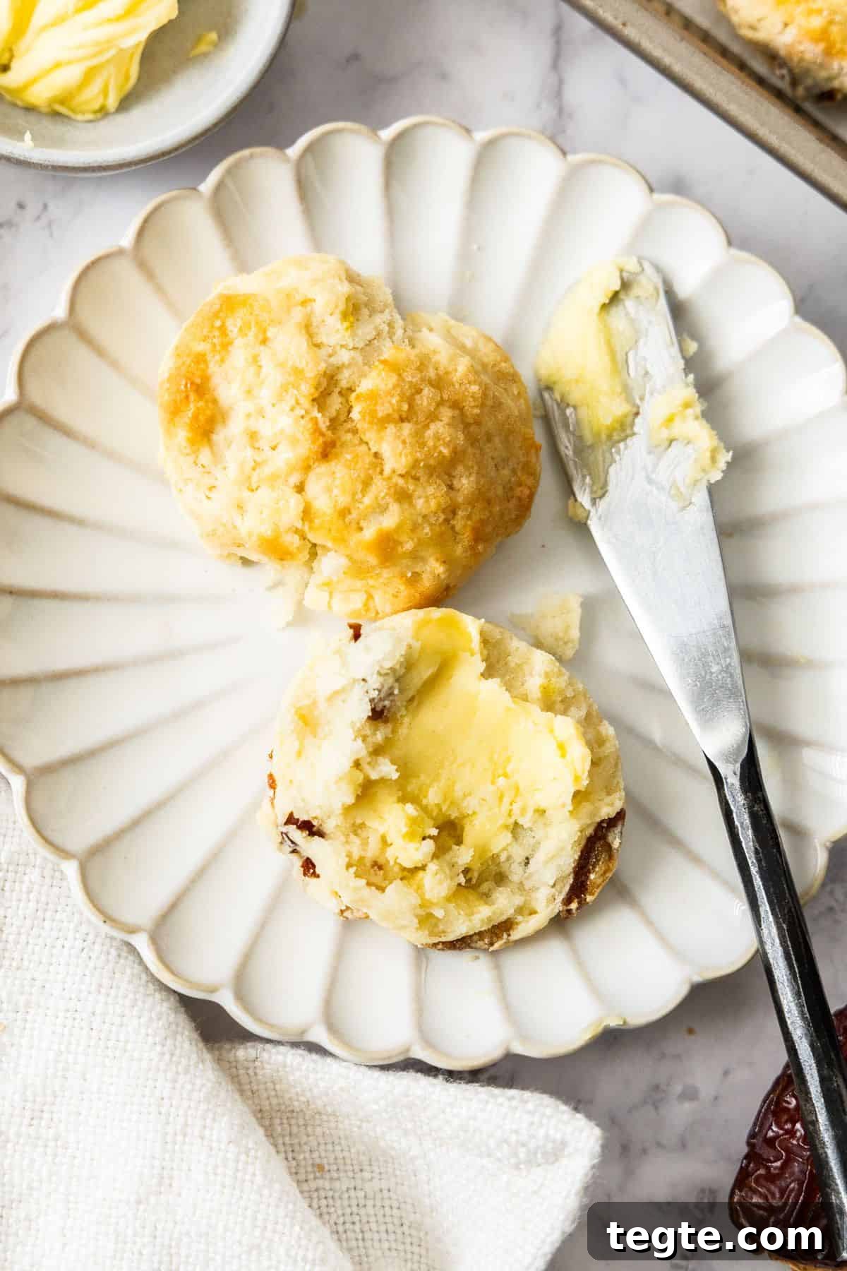 Chewy Date and Zesty Lemon Scones Recipe 8 Round plate with a Date and Lemon Scone cut in half, with a knife and some butter on the edge.