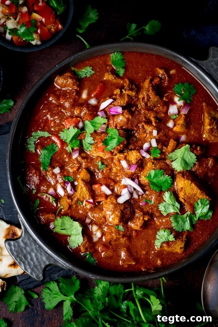 Overhead image of beef curry in a dark dash on a dark background, topped with coriander, chillies and red onion