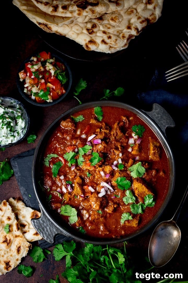 Overhead of slow cooked beef curry in a dish on a dark background with ingredients scattered around