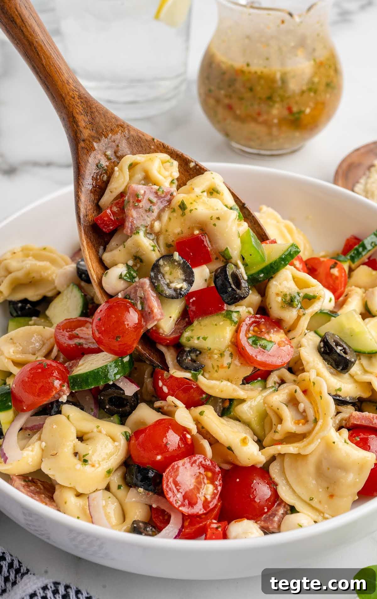 Wooden spoon lifting a serving of tortellini pasta salad featuring tomatoes, cucumbers, olives, and salami.