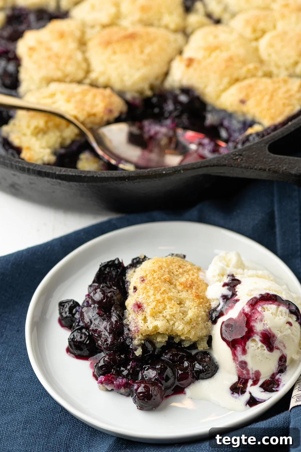 Low-Carb Blueberry Cobbler (Gluten-Free, Sticky Sweet) 4 A serving of low carb blueberry cobbler on a white plate with a scoop of melting vanilla ice cream with blueberry drizzle . A cast iron pan in the background.
