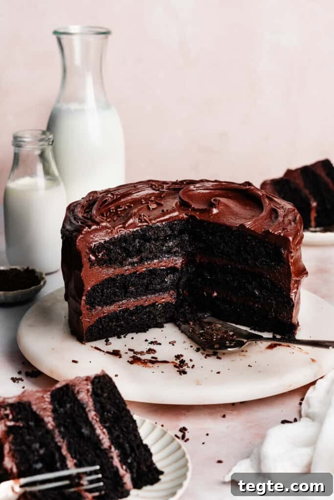 Eggless Chocolate Cake with Decadent Chocolate Fudge Frosting 2 The sliced eggless chocolate cake on a marble cake plate, with slices of cake and glasses of milk nearby.