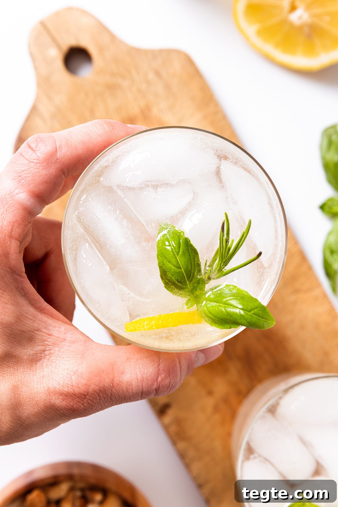 Glass filled with rosemary basil gin fizz with hand holding the glass with lemon wedge and herbs in glass with wood board underneath.