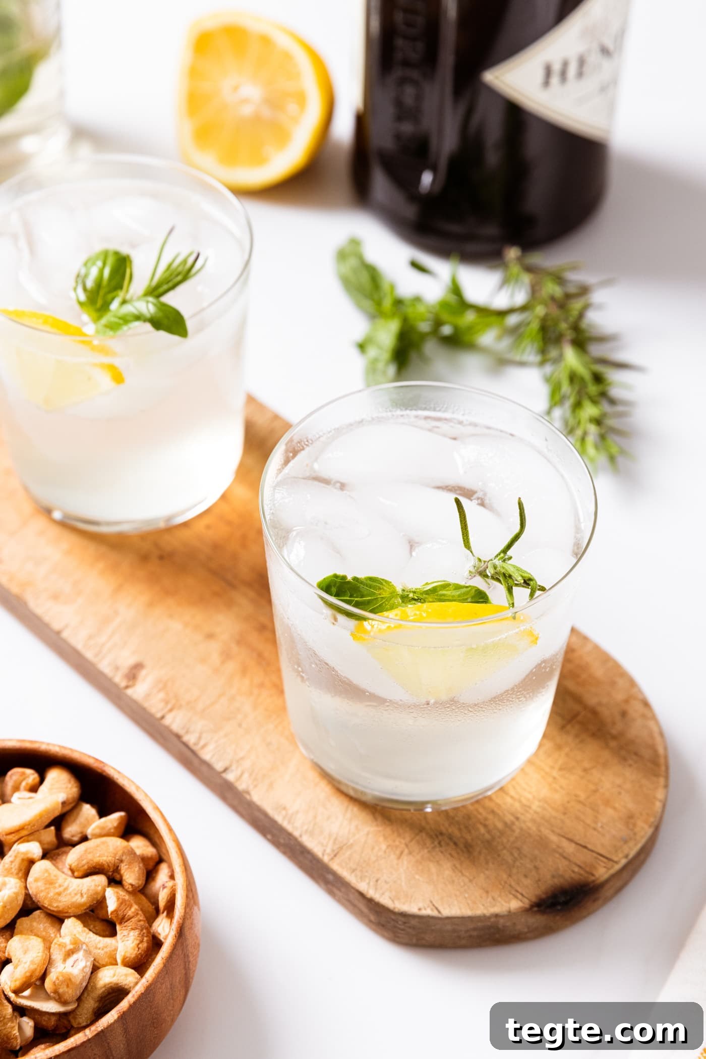 Two glasses filled with clear gin fizz liquid with lemon wedges and herbs stuck in glass sitting on wood board with small bowl of peanuts beside and alcohol container in background.