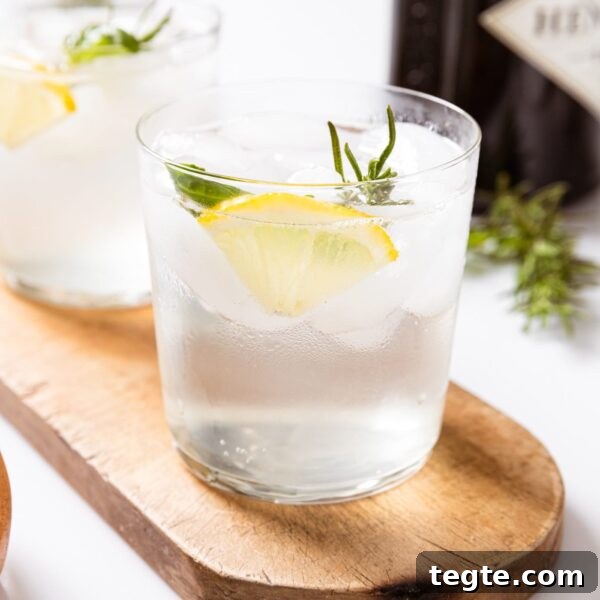 Two glasses filled with clear gin fizz liquid with lemon wedge and herbs sitting in glass sitting on wood board with small bowl filled with nuts.