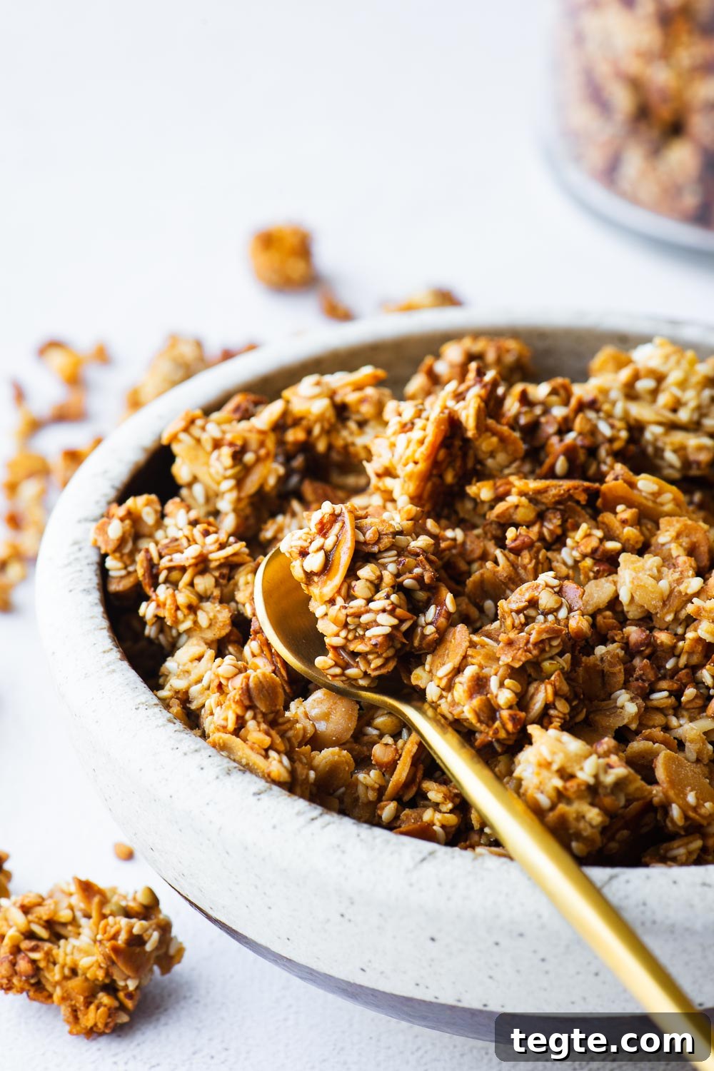 Close-up of tahini granola with sesame seeds in a ceramic bowl with a jar in the background and a small gold spoon.