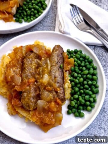 A plate of sausages on a bed of sweet potato mash covered in tomato and onion gravy with a side helping of peas.