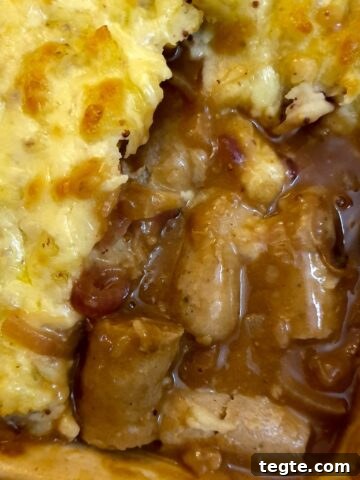 Pieces of sausages with onion gravy in the corner of a casserole dish, surrounded with cheesy mashed potatoes.