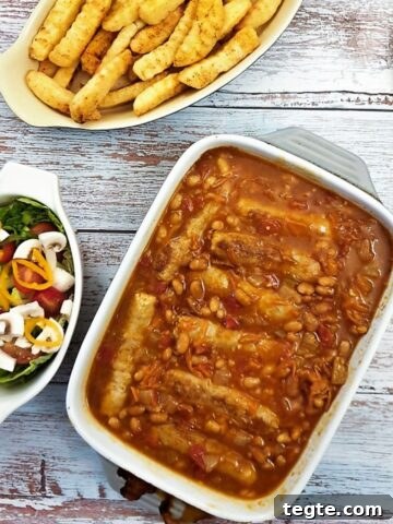 A dish of sausage and bean casserole on a table alongside dish of crispy french fries and a green salad.